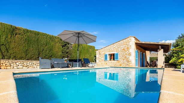 A clear swimming pool with lounge chairs and an umbrella, surrounded by stone walls and a stone building with blue shutters.