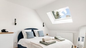 A bedroom with a sloped ceiling, a skylight, a bed with pillows, a bedside table, and a mirror. - Gîtes de France® - Domaine Majorelle (LA BELLIERE)