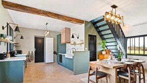 A modern kitchen with a central island, a dining area with a wooden table, and a staircase leading to an upper level. - Gîtes de France® - Domaine Majorelle (LA BELLIERE)