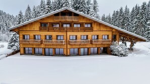 Eine Holzchalet mit mehreren Balkonen, großen Fenstern und einem geneigten Dach, umgeben von schneebedeckten Bäumen.