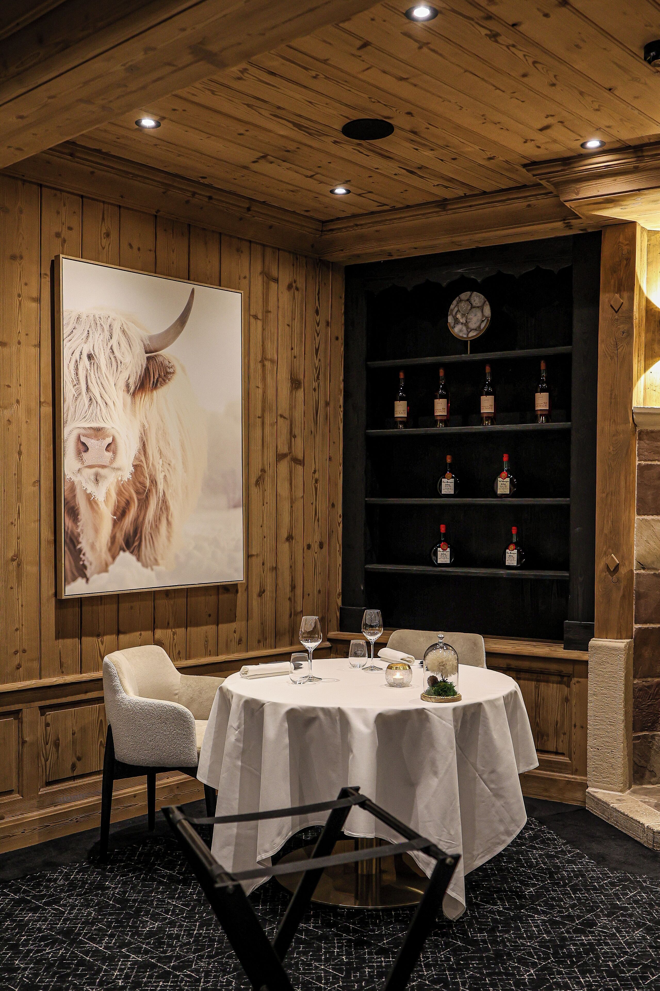 Un comedor con una mesa redonda, sillas, un cuadro grande enmarcado de una vaca y una estantería para vinos con botellas.