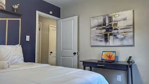 Habitación de hotel con cama, escritorio, computadora portátil, puerta y un cuadro abstracto colgado en la pared.