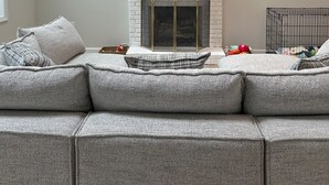A modern living room with a white brick fireplace, a flat-screen TV mounted above it, and a large sectional sofa with grey cushions. - Your Wolfeboro Home Away From Home (Wolfeboro)