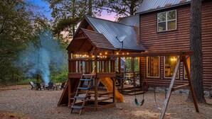 A wooden cabin with a playhouse, a swing set, and a dining area with outdoor seating.