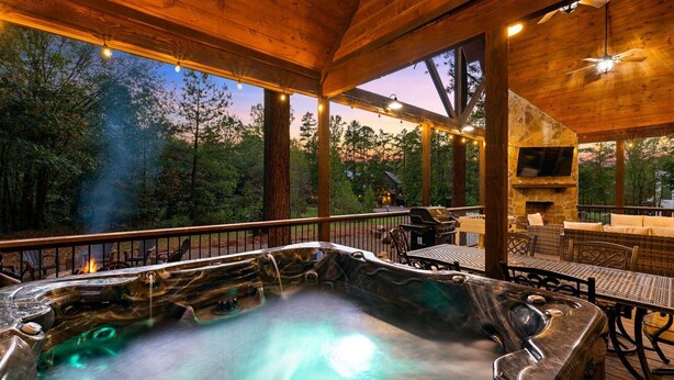A hot tub with a stone surround, wooden deck, outdoor seating, and a stone fireplace.