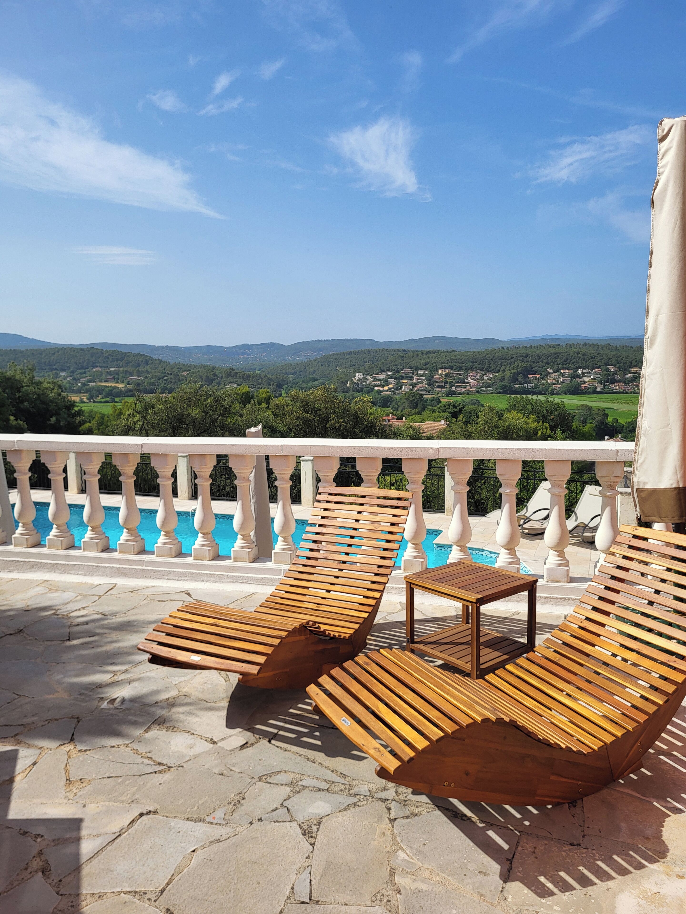 A balcony with wooden lounge chairs and a small table, overlooking a pool and a scenic view of hills and a village.