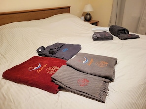 A neatly made bed with a white comforter, a lamp on the nightstand, and folded towels and robes on the bed.