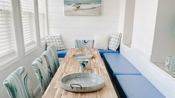Une longue table à manger en bois, assortie de chaises, un banc bleu avec coussin et, au mur, un tableau représentant une scène de plage.
