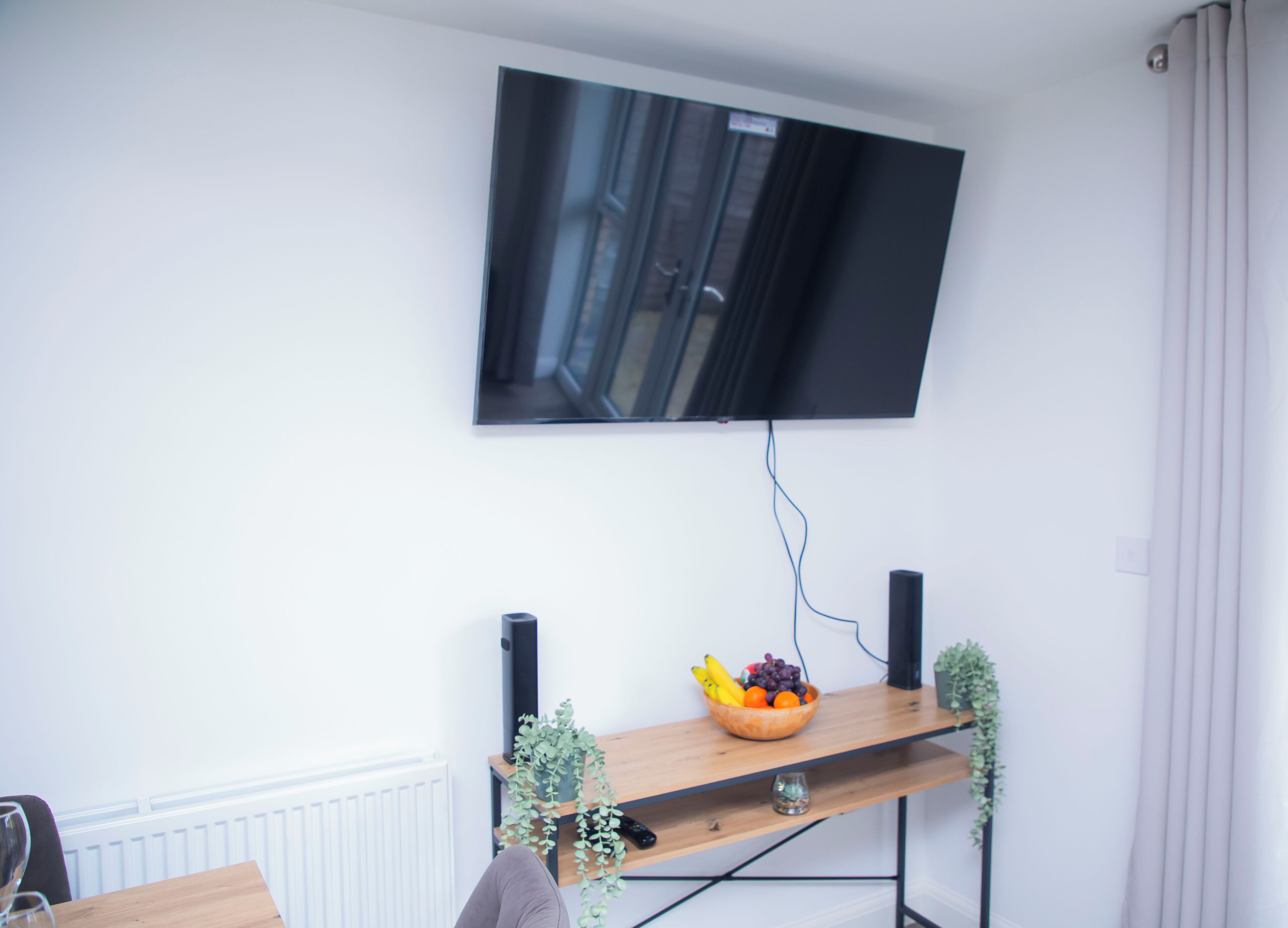 Uma sala de estar moderna com uma televisão de tela plana montada, uma estante de madeira com uma fruteira, caixas de som e um vaso com planta.