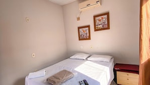 Ein Zimmer mit einem Einzelbett, einer weißen Bettdecke, einer Fernbedienung und einem beigen Handtuch auf dem Bett. An der Wand hängen zwei gerahmte Kunstwerke, ein Deckenventilator und eine Klimaanlage.
