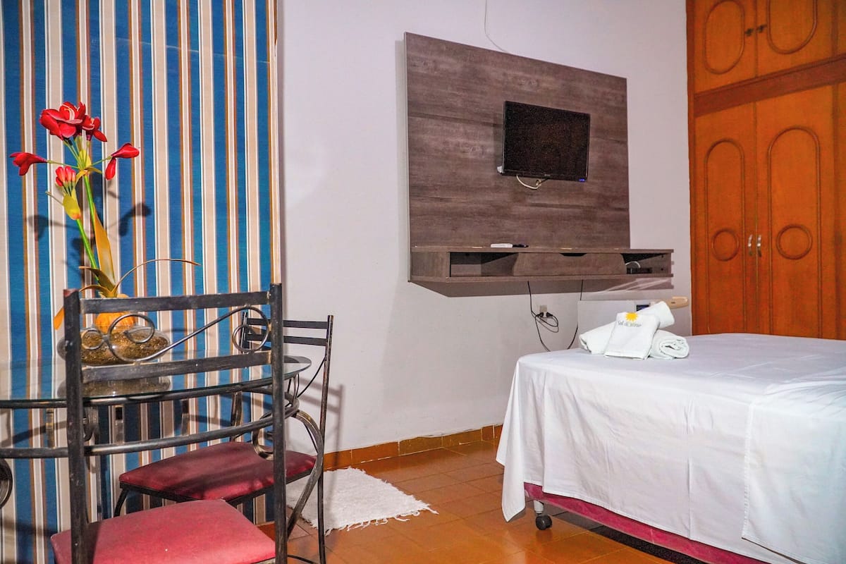 Quarto de hotel com cama, televisão fixada na parede, uma cadeira e uma mesinha com um vaso de flores.