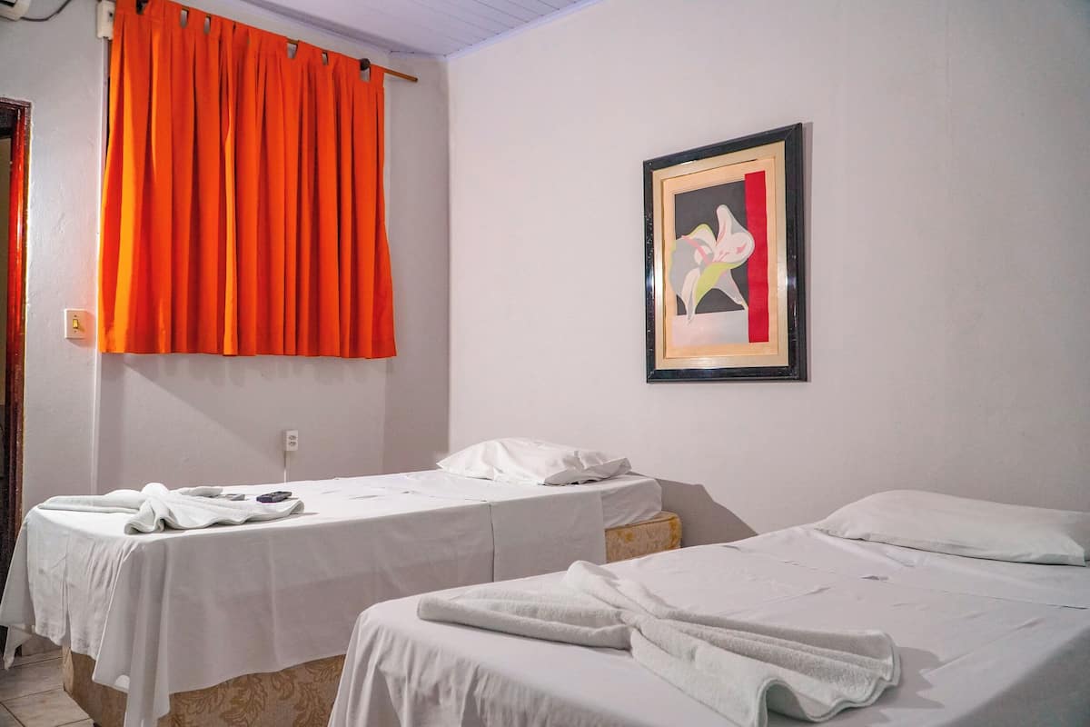 Duas camas de solteiro com lençóis brancos, uma cortina laranja vibrante e um quadro abstrato na parede.