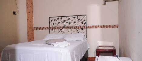 A neatly made bed with white linens, a headboard with a leaf pattern, a small bedside table with a lamp, and a wall-mounted shelf with flowers.