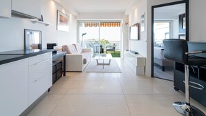 Ein moderner, gut beleuchteter Wohnbereich mit Sofa, Couchtisch und Esstisch. Es gibt eine Küchenzeile mit Schränken und einer Arbeitsplatte. Das Zimmer verfügt über ein großes Fenster mit Blick ins Freie.
