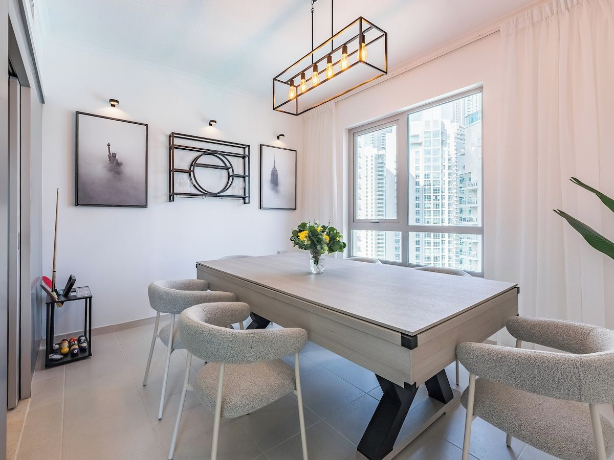 Uma sala de jantar moderna com uma mesa grande, quatro cadeiras e um vaso de flores. Há quadros nas paredes, uma janela com vista para prédios e um lustre.