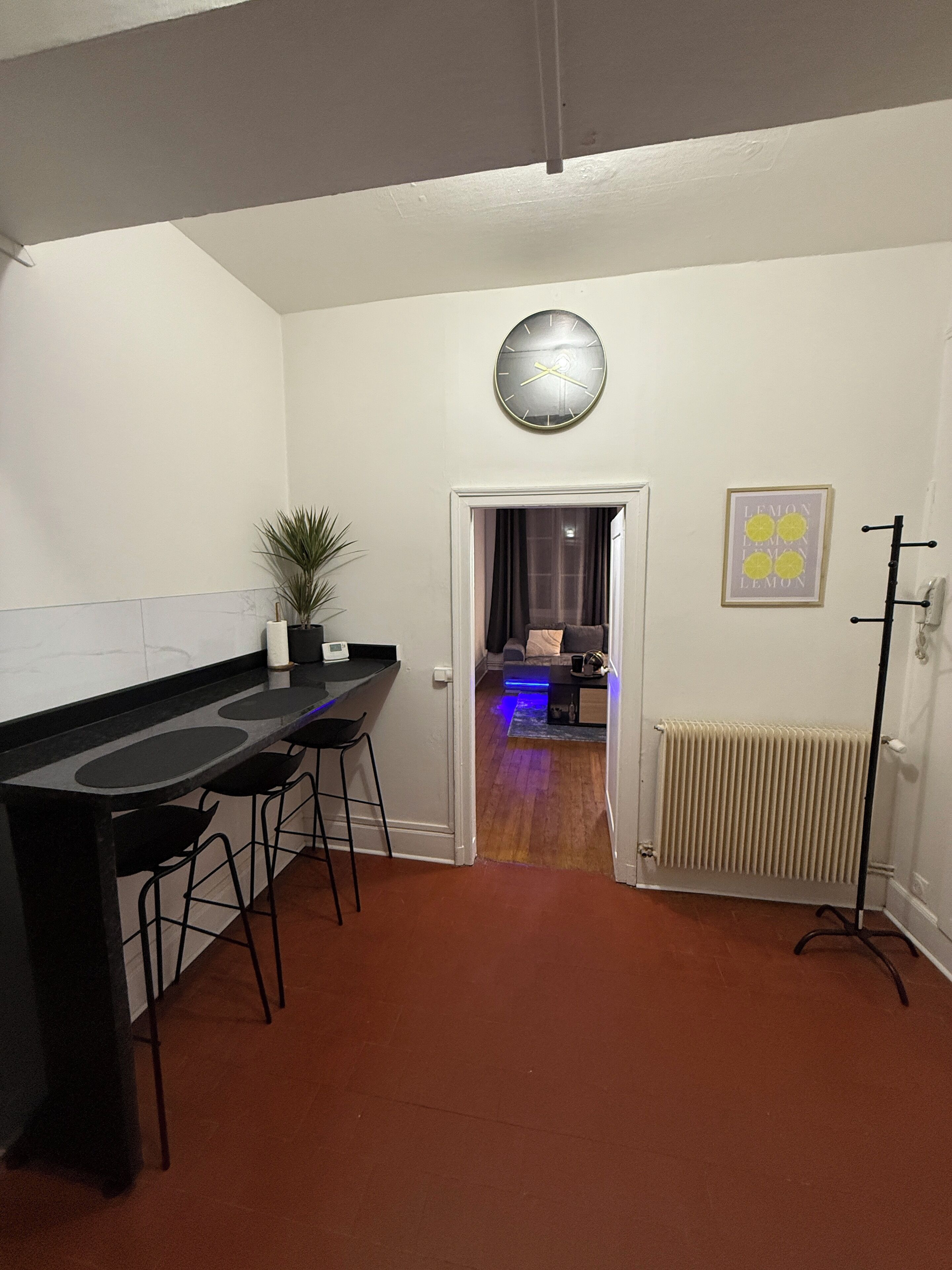 Et køkken med en sort bordplade, to barstole, en potteplante, et vægur og en radiator.