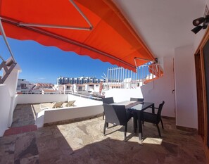 A rooftop terrace with a table and chairs, a large orange umbrella, and a view of buildings in the background.
