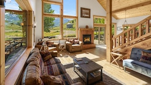 A spacious living room with a fireplace, large windows, and a wooden staircase.