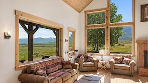 A cozy living room with large windows offering a view of a green landscape and mountains. There are two sofas and a coffee table.