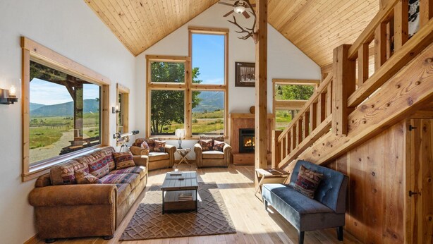 A cozy living room with a fireplace, large windows, and a wooden staircase.