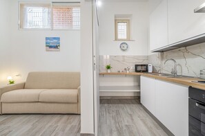 A modern kitchen with a microwave, sink, and cabinets, a beige sofa, and a small window with patterned curtains.