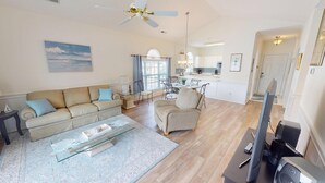 A spacious living room with a beige sofa, a glass coffee table, and a dining area with a white table and chairs. A kitchen with white cabinets and a dining area with a chandelier is visible in the background.