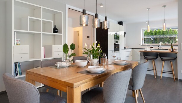 Un comedor moderno con una mesa de madera, sillas grises, luces colgantes y vista a la cocina.