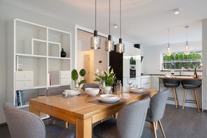 Un comedor moderno con una mesa de madera, sillas grises, luces colgantes y vista a la cocina.