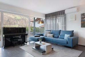 Un salon moderne avec un piano, un canapé bleu, une table basse et une grande fenêtre donnant sur des collines.