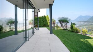 Un balcón moderno con puertas de vidrio, suelo de concreto y vista a un paisaje verde con árboles y montañas.