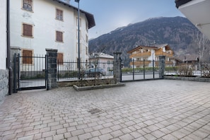 A paved courtyard with a stone fence, a building with wooden shutters, and a mountain in the background.
