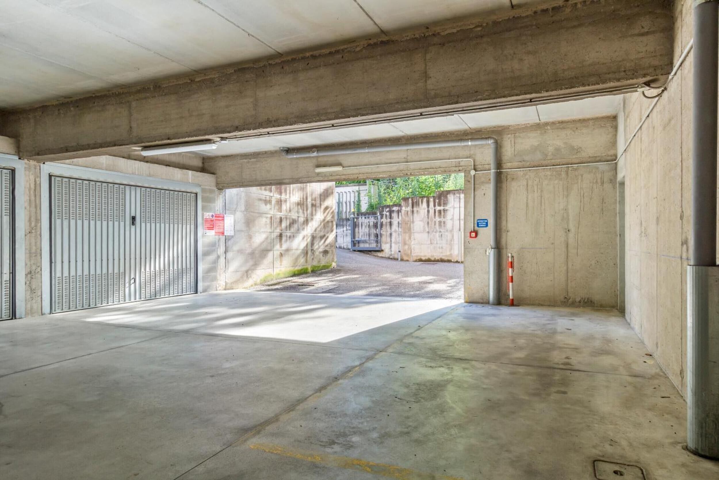 Un parking en béton, doté de portails métalliques, d’un extincteur et d’un panneau bleu indiquant une zone de stationnement.
