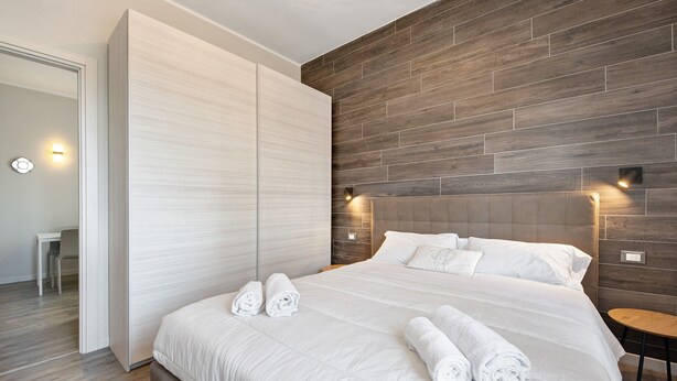 A modern bedroom with a large bed, wooden headboard, and two wall-mounted lights. - Apartment 'Garda Flow' with Wi-Fi and Air Conditioning (Lonato del Garda)