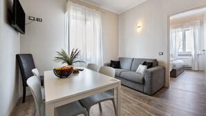 A modern living room with a sofa, a dining table with chairs, a TV mounted on the wall, and a window with curtains. - Apartment 'Garda Flow' with Wi-Fi and Air Conditioning (Lonato del Garda)