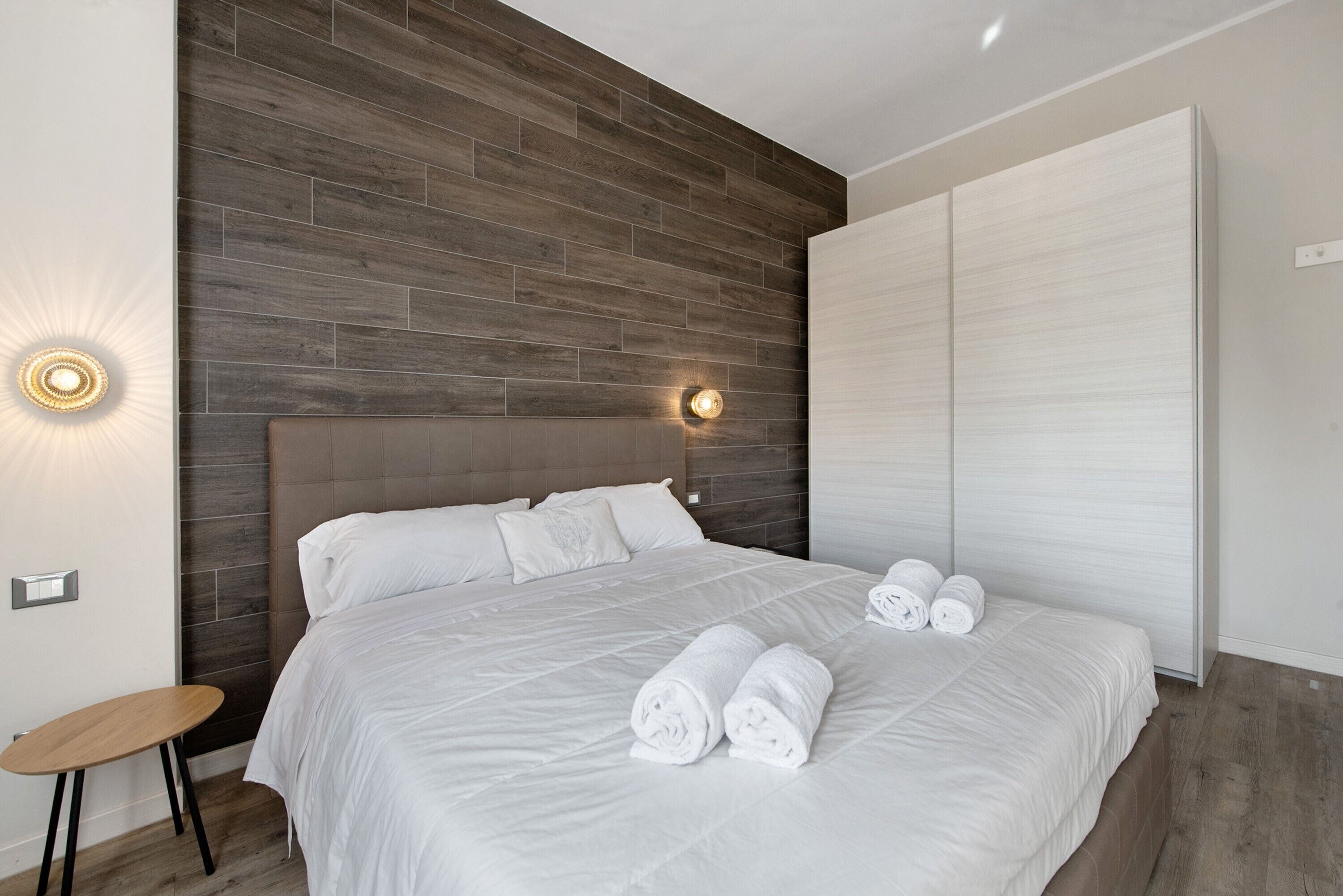 Una habitación de hotel moderna con una cama grande, un panel decorativo de madera, una lámpara fijada a la pared y un armario espacioso.