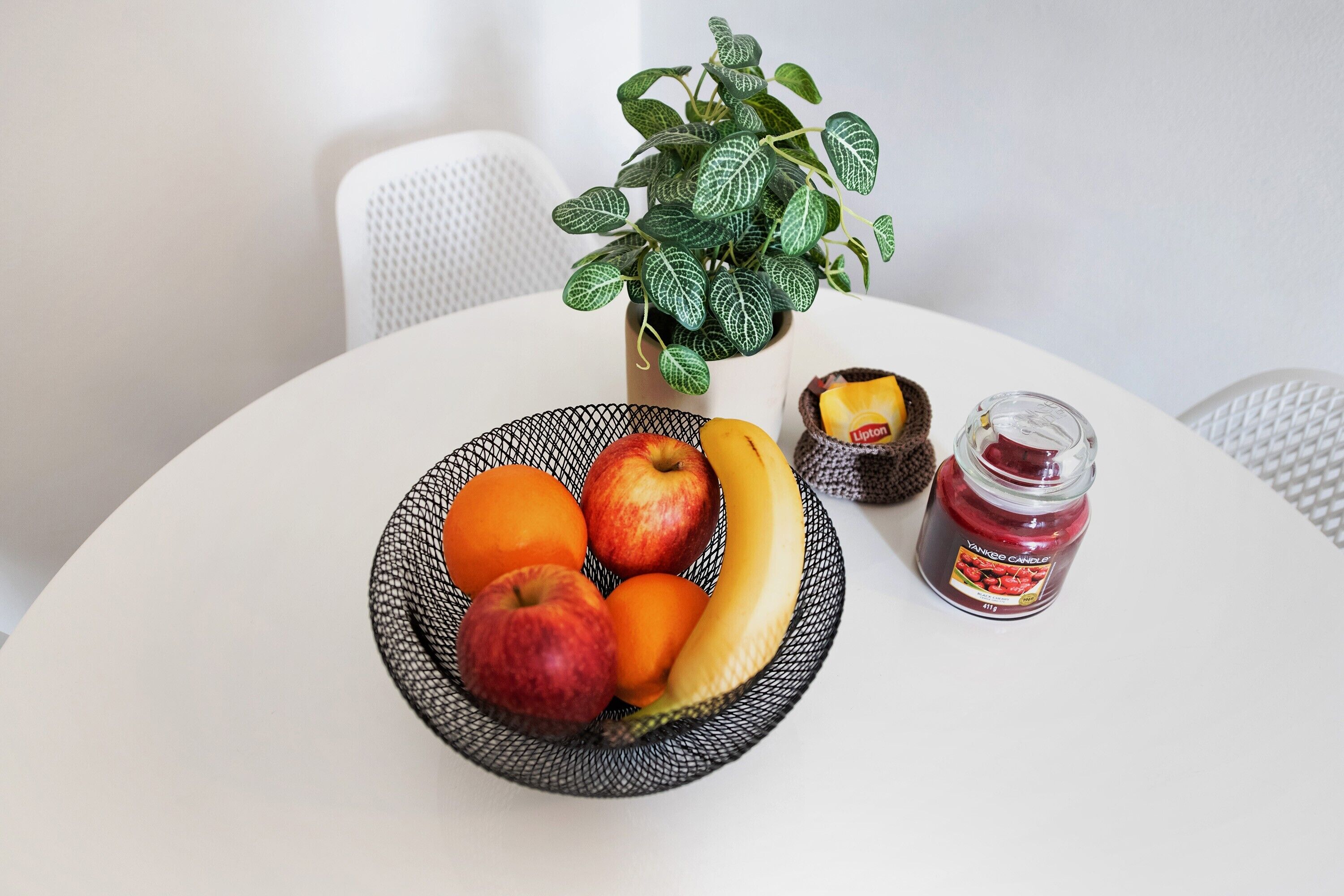A table with a fruit bowl containing apples, oranges, and a banana, a potted plant, and a jar of jam.