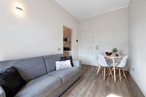 A modern living room with a grey sofa, white cushions, a dining table with white chairs, and a small kitchen visible through an open door. - Apartment 'Garda Flow' with Wi-Fi and Air Conditioning (Lonato del Garda)