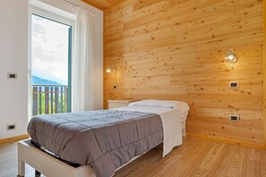 A bedroom with a wooden wall, a bed with a grey bedspread, a white bedside table, and a window with a view of greenery.