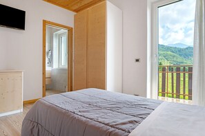 A bedroom with a bed, a wooden wardrobe, a television, and a balcony with a view of greenery. - Apartment 'Montibeller Cima 12' with Mountain View, Shared Garden and Wi-Fi (Roncegno Terme)