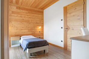 Une chambre avec un plafond en bois, un lit recouvert d’une couverture grise, une table de chevet et une porte en bois.