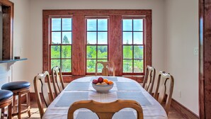 Comedor con una mesa larga, sillas de madera, un bol de fruta y ventanas amplias con vistas a la vegetación.