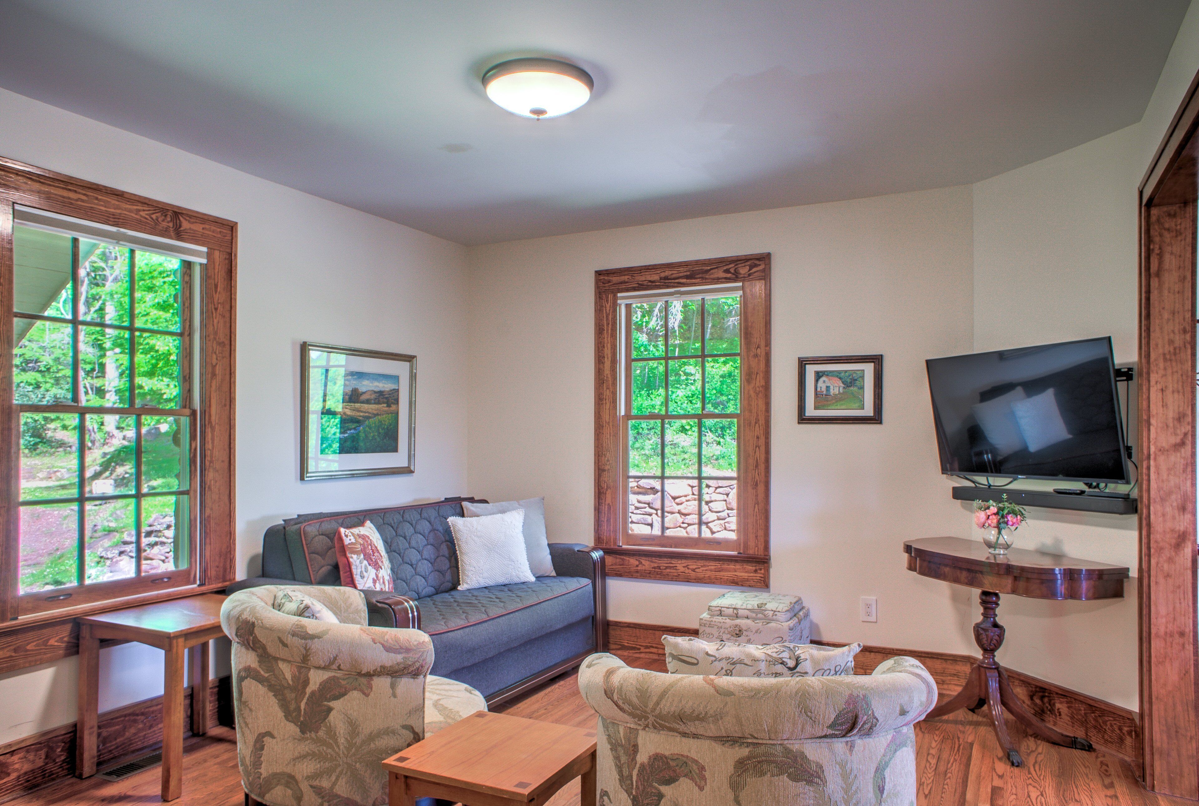 A living room with a sofa, two armchairs, a coffee table, a TV mounted on the wall, and large windows with a view of greenery.
