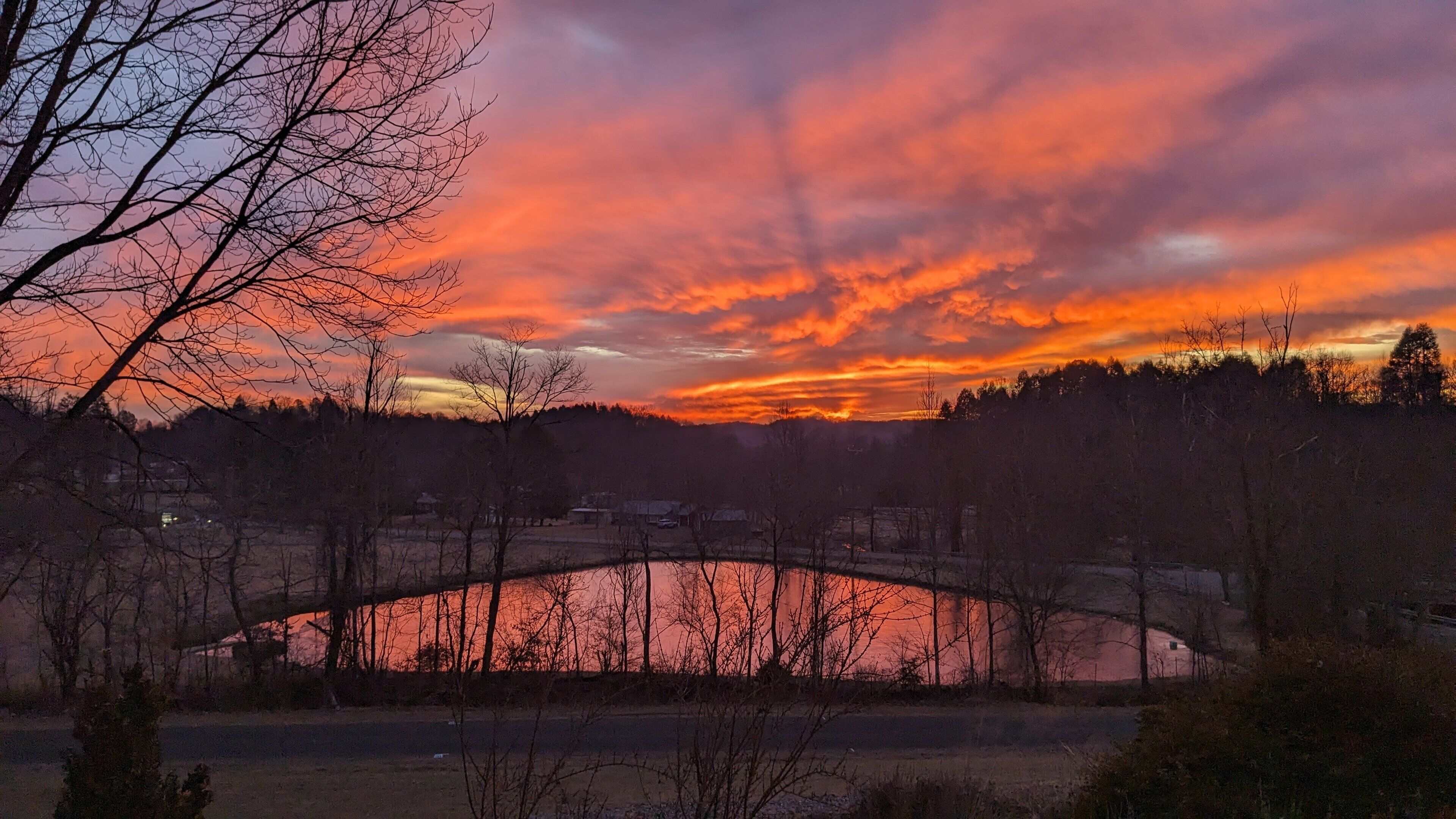 A sunset over a bridge with a river below, surrounded by trees and a few buildings in the distance.