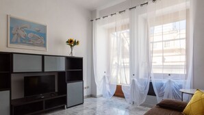 Ein Wohnzimmer mit Fernseher, einem Gemälde, einer Blumenvase, einer Couch und durchscheinenden Vorhängen vor einem Fenster.
