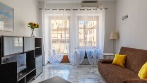 Ein modernes Wohnzimmer mit einer Couch, einem Fernseher und Blick auf Gebäude durch transparente Vorhänge.