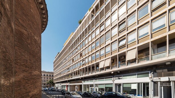 Ein mehrstöckiges Gebäude mit einer rasterförmigen Fassade, großen Fenstern und einem Parkplatz davor.