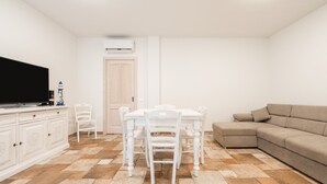 A living room with a television, a sofa, a dining table with chairs, and a door. - Holiday Home 'Giardino' with Private Garden, Wi-Fi and Air Conditioning (Villasimius)