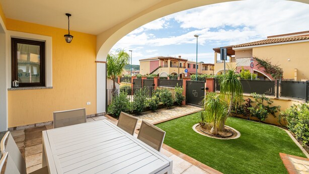A patio with a table and chairs, a small lawn, and a view of neighboring buildings. - Holiday Home 'Giardino' with Private Garden, Wi-Fi and Air Conditioning (Villasimius)