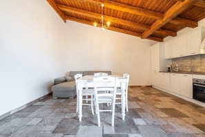 A kitchen with white cabinets, a dining table with chairs, a grey sofa, and a wooden ceiling.