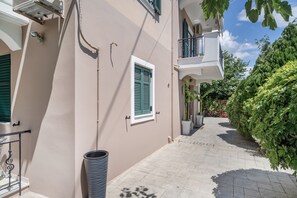 A narrow pathway leading to a building with a balcony, a window with shutters, and a trash bin.